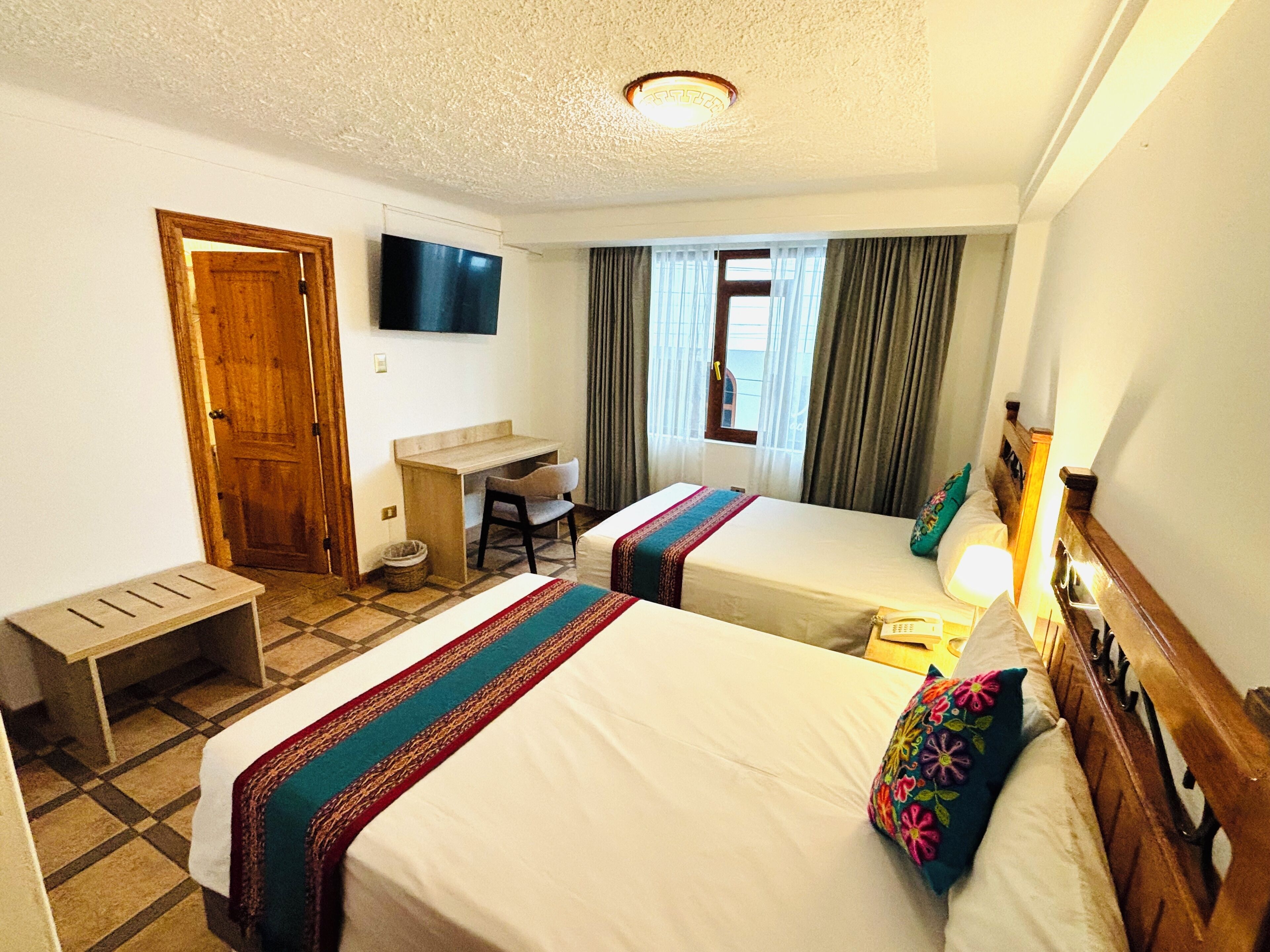 A hotel room with two beds, a desk, a TV, and a window with curtains.