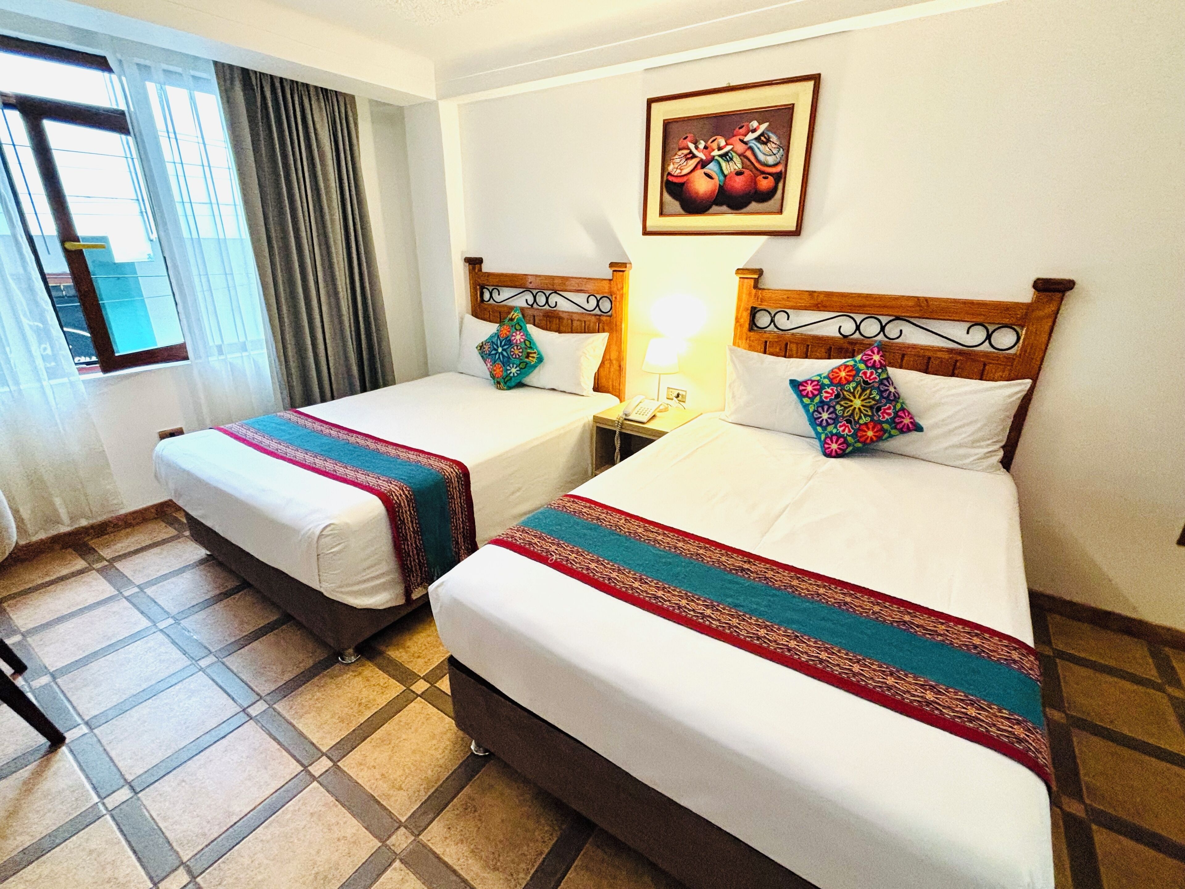A hotel room with two beds, a window with curtains, a painting on the wall, and a tiled floor.