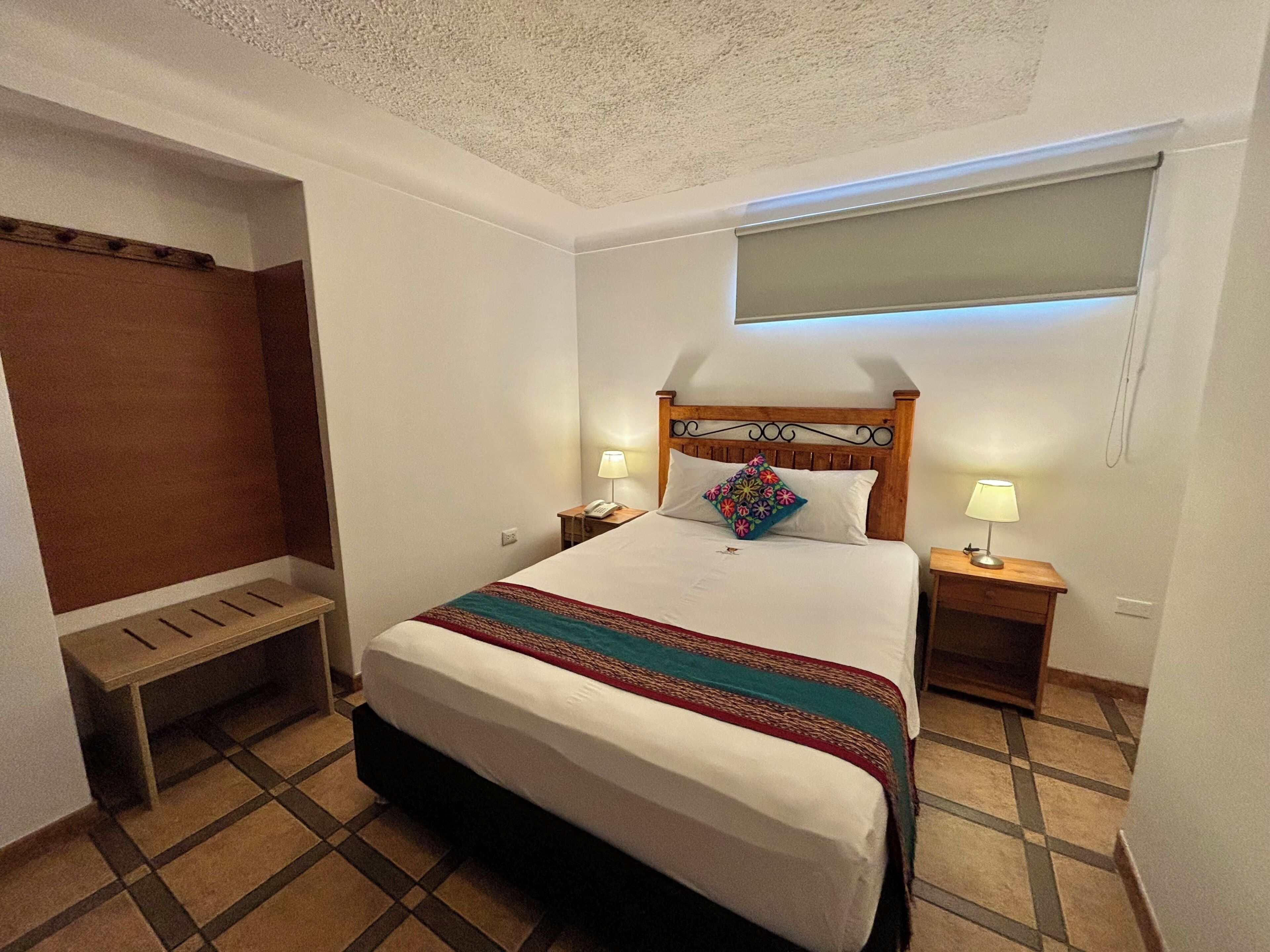 Habitación de hotel con cama, dos mesitas de noche con lámparas, cabecero de madera, ventana con persianas, un banco y un armario.