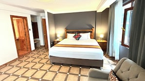 A hotel room with a large bed, a bedside table with a lamp, a chair, a wooden door, and a checkered floor. - Hotel Rojas Machupicchu (Aguas Calientes)