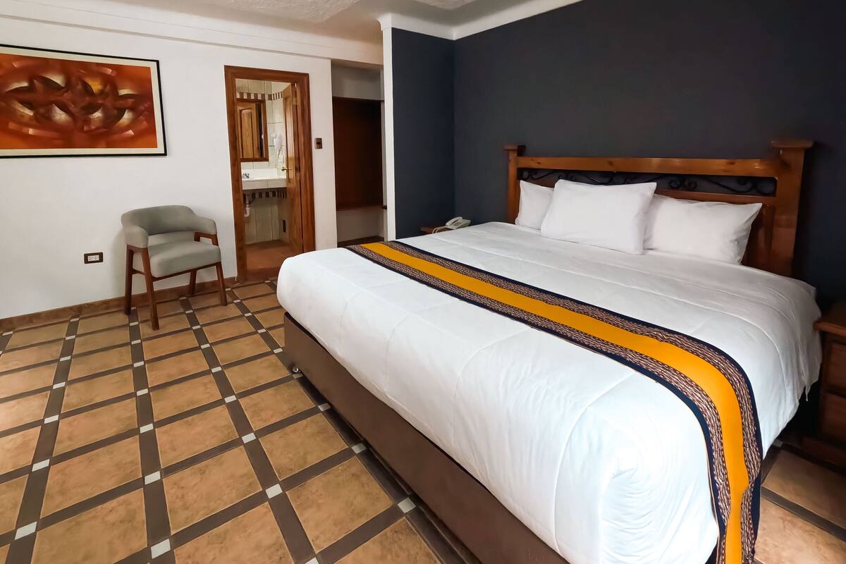 A hotel room with a large bed, a chair, a bathroom with a mirror, and a wall with a painting.
