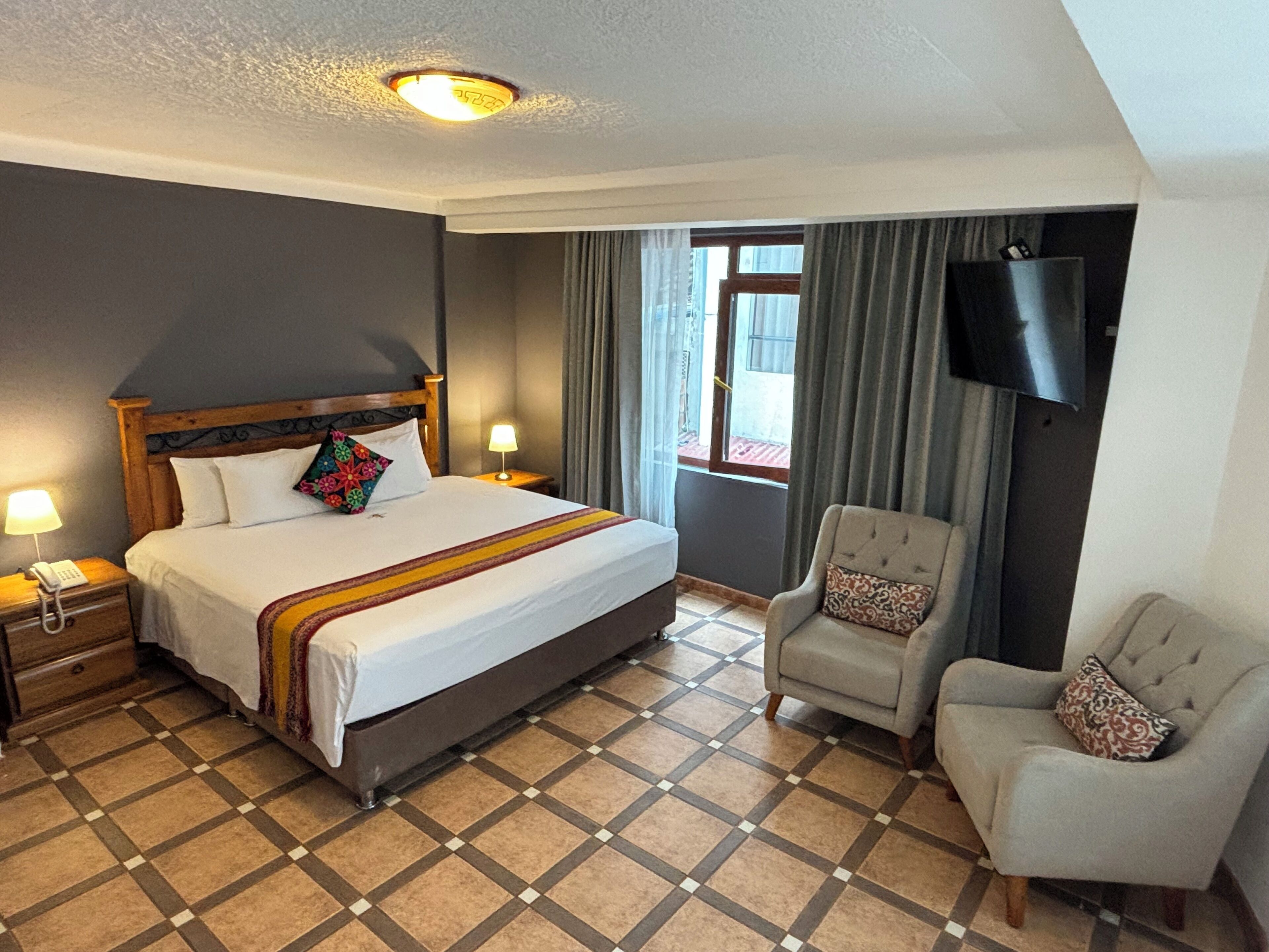 Une chambre d’hôtel avec un grand lit, une chaise, un téléviseur fixé au mur et une fenêtre avec des rideaux.