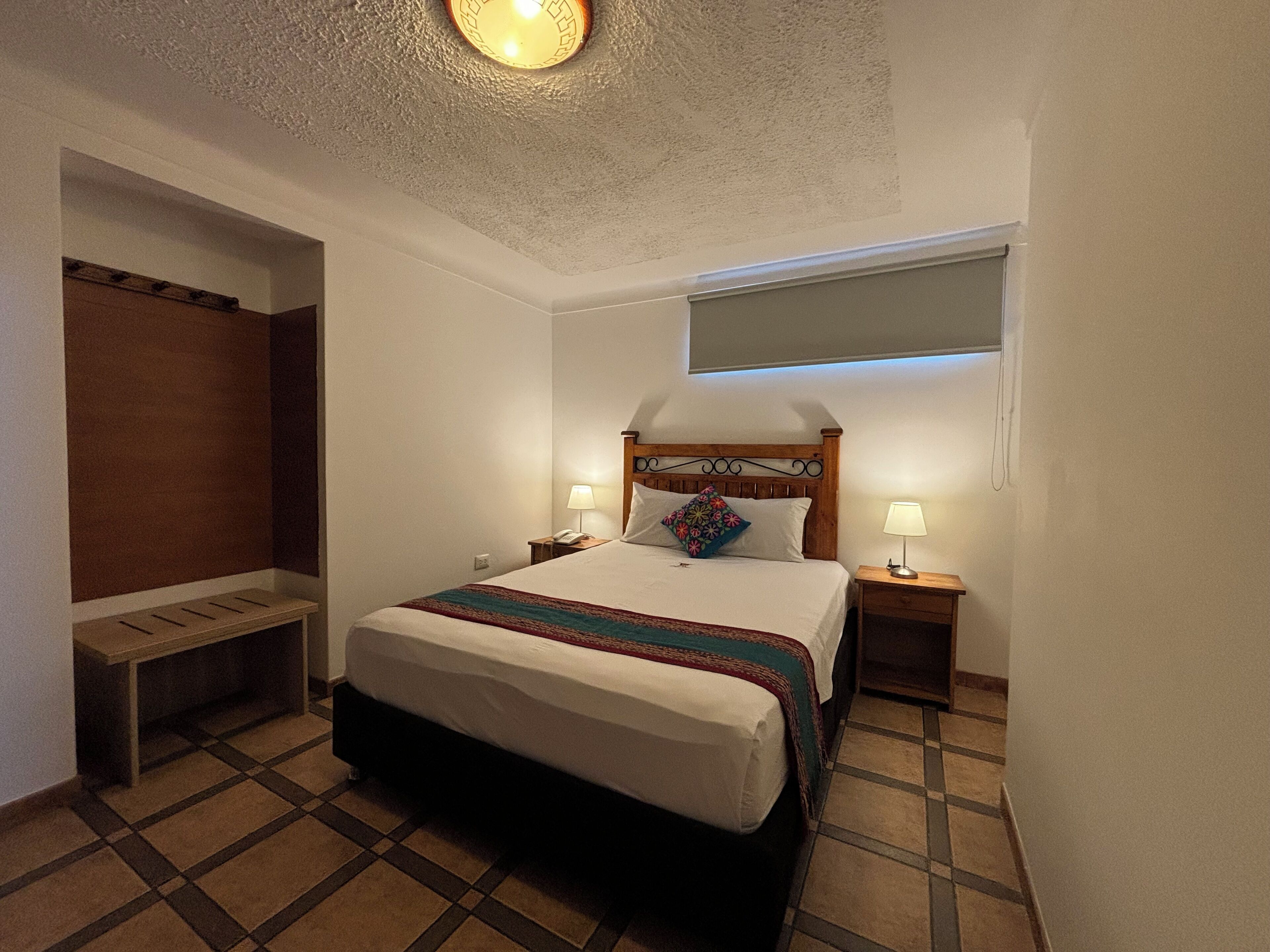 Una habitación de hotel con cama, lámparas de noche, un banco y una lámpara de techo.