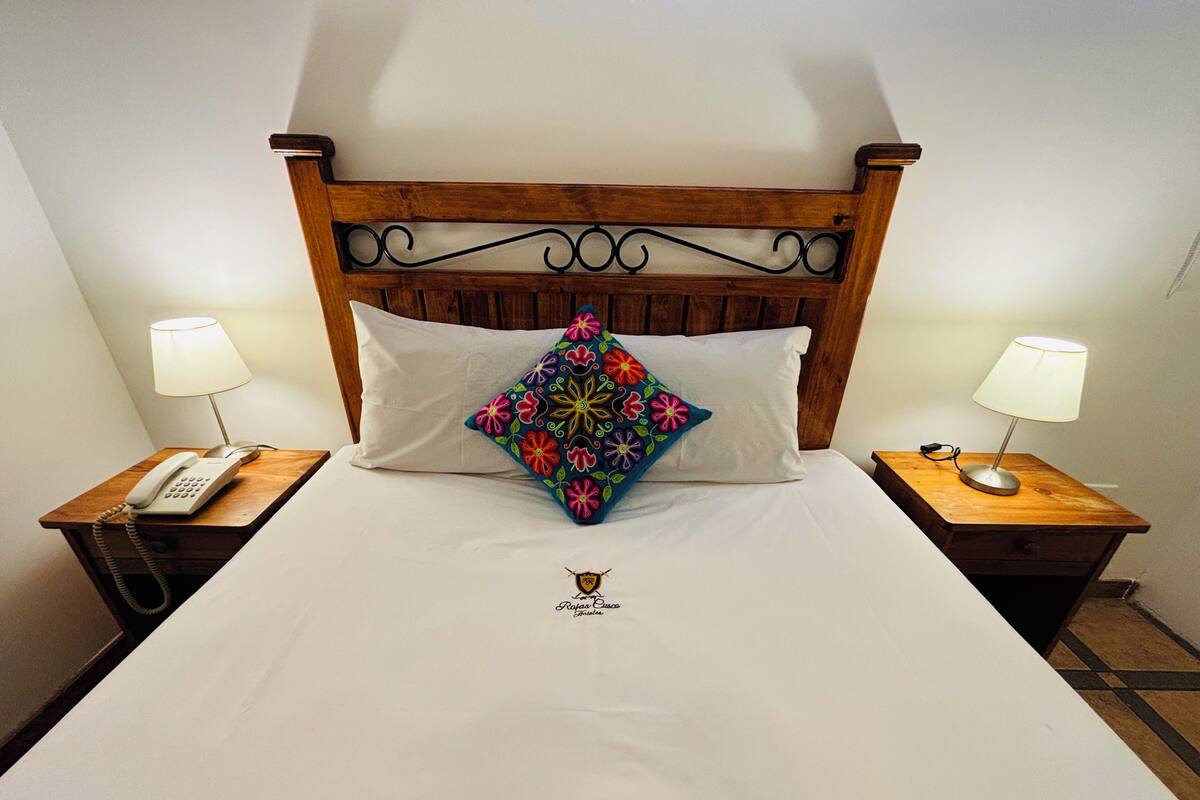 A hotel room with a wooden headboard, a white bedspread, a decorative pillow, two bedside tables with lamps, and a telephone on one table.