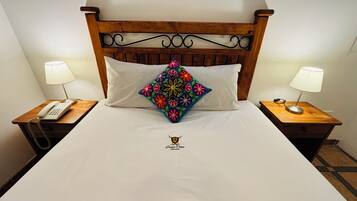 Habitación de hotel con cabecera de madera, colcha blanca, almohada decorativa, dos mesitas de noche con lámparas y un teléfono sobre una de ellas.