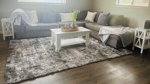 Ein Wohnzimmer mit grauem Sofa, weißem Couchtisch und grauem Teppich. Zwei Fenster mit Jalousien und ein Nachttisch mit Lampe sind ebenfalls zu sehen.