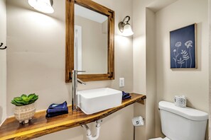 Une salle de bain avec un meuble-lavabo en bois, un lavabo blanc, un miroir avec un cadre en bois, un luminaire fixé au mur, une plante en pot et des toilettes.