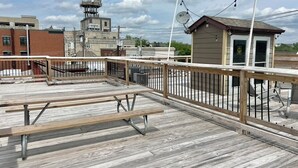 A wooden deck with picnic tables and a metal railing, overlooking a cityscape with buildings and a tower. - Downtown studio apartment with ROOFTOP DECK (Bloomington)