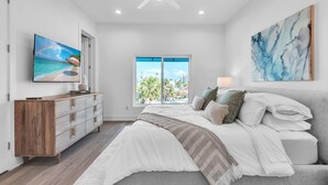 Ein Schlafzimmer mit Bett, Schminktisch, Fernseher, Deckenventilator und einem Fenster mit Blick auf Palmen.