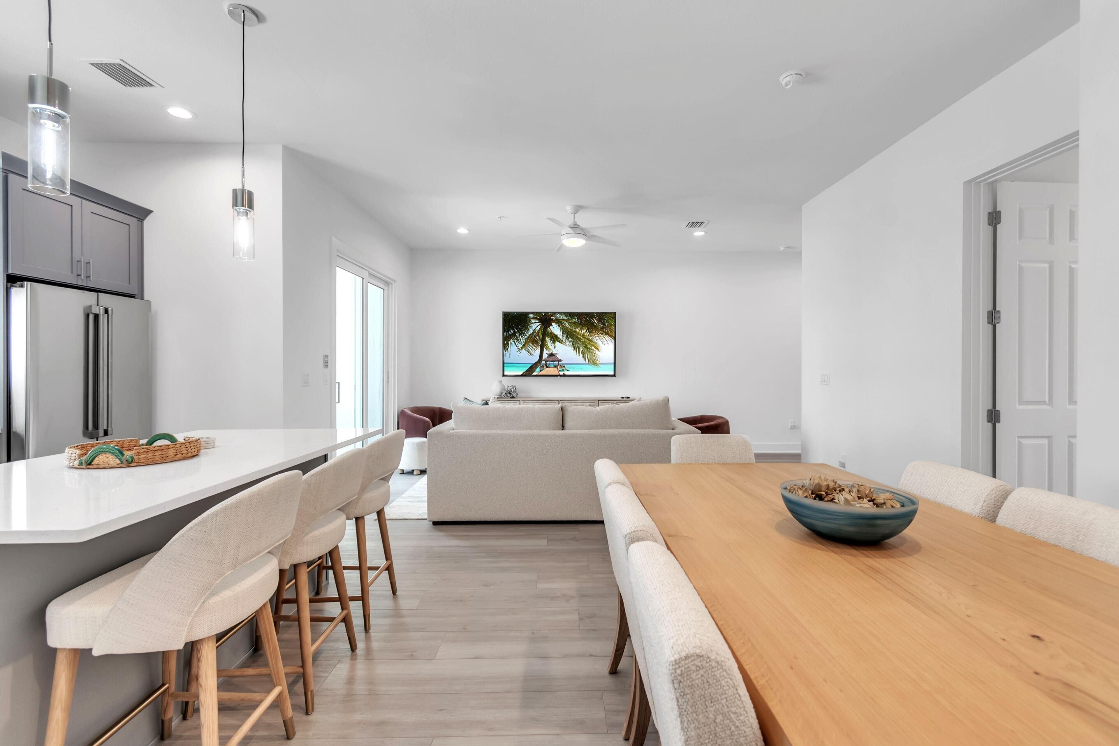 Une cuisine moderne avec un îlot central, des tabourets de bar, une table à manger en bois et un grand tableau représentant une scène de plage.