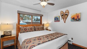 Habitación de hotel con cama, ventana, ventilador de techo, mesita de noche y decoración en la pared.