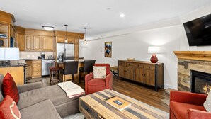 A modern living room with a fireplace, wooden furniture, and a kitchen area with a refrigerator and dining table.