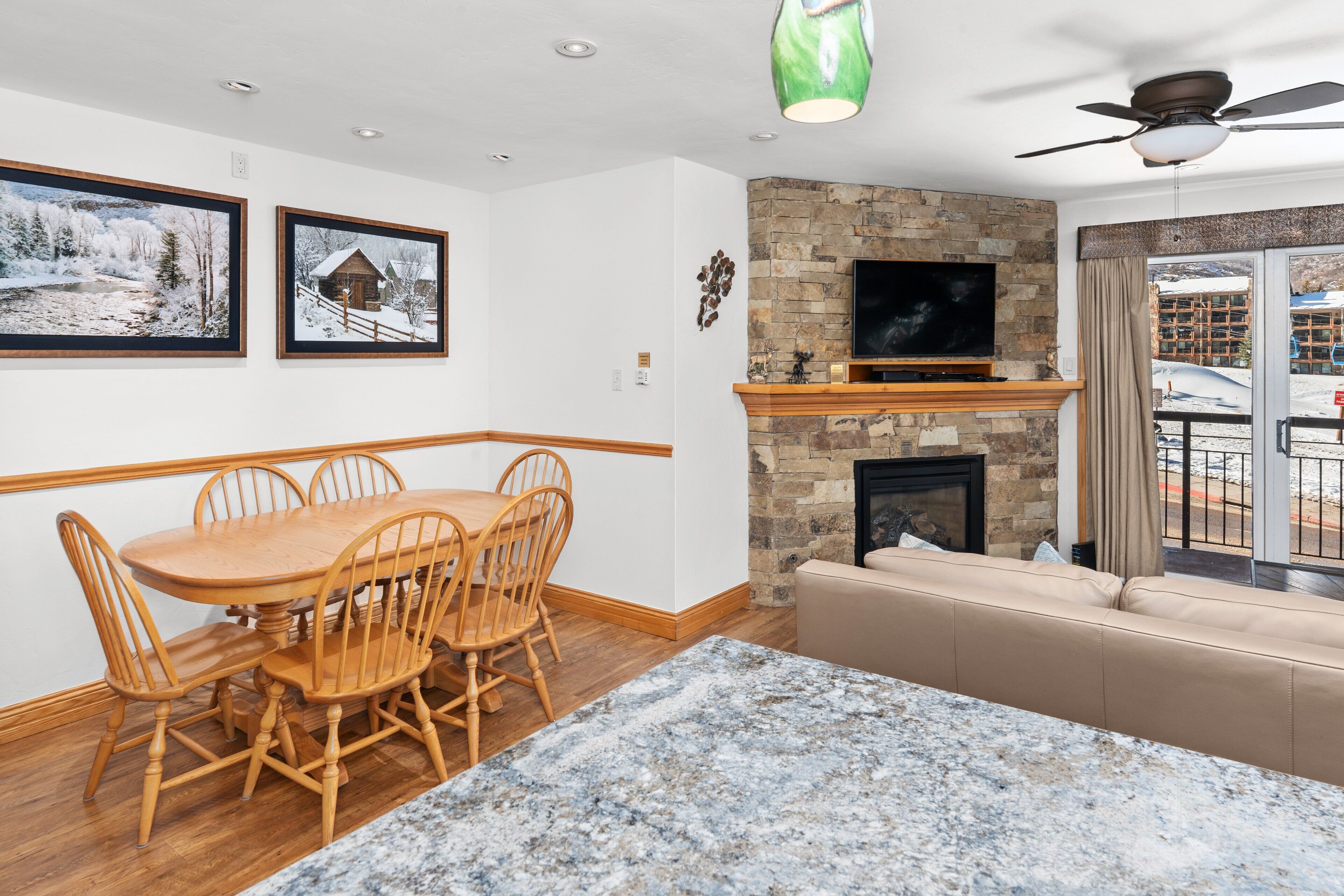 A dining area with a wooden table and chairs, a stone fireplace, a television, and a balcony with a view.
