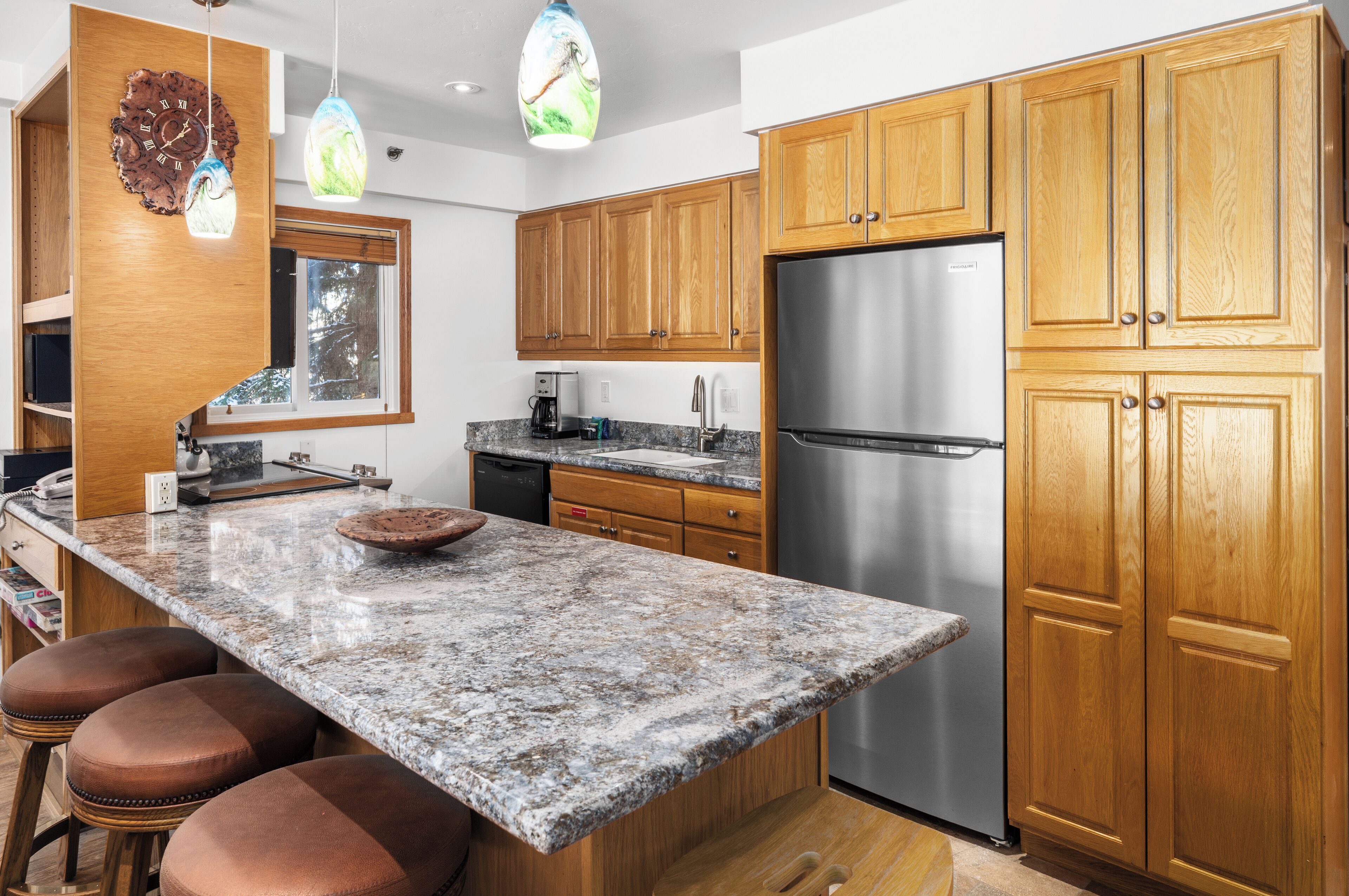 A modern kitchen with wooden cabinets, a stainless steel refrigerator, granite countertops, and pendant lighting.