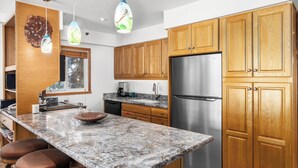 A modern kitchen with wooden cabinets, a stainless steel refrigerator, granite countertops, and pendant lighting. - Crestwood 1202 Deluxe 3 Bedroom (Snowmass Village)