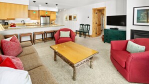 A modern living room with a wooden coffee table, red sofas, and a kitchen area with wooden cabinets and a stainless steel refrigerator.