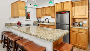 A kitchen with wooden cabinets, a granite countertop, a stainless steel refrigerator, and a bar area with stools.