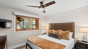 A bedroom with a bed, a ceiling fan, a TV mounted on the wall, a window with a view of trees, and a bedside table with a lamp.