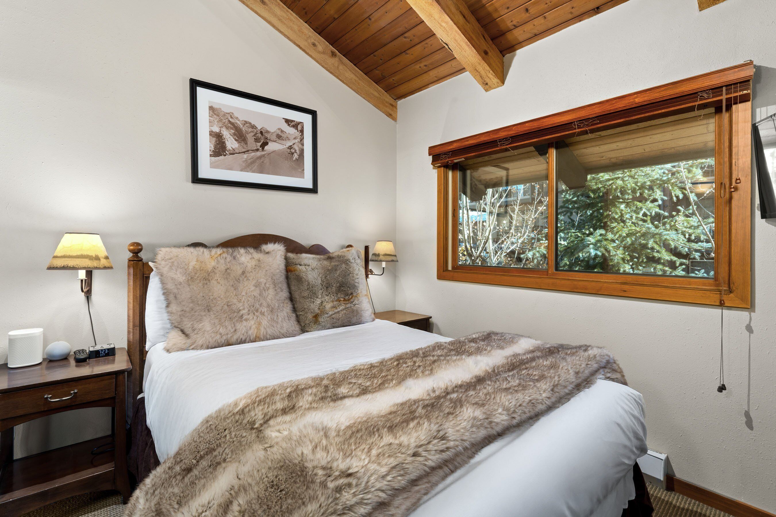 A bedroom with a wooden headboard, a large window with a view of greenery, a framed picture on the wall, and a bedside table with a lamp and a radio.