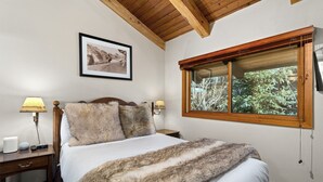 A bedroom with a wooden headboard, a large window with a view of greenery, a framed picture on the wall, and a bedside table with a lamp and a radio. - Crestwood 1207 Deluxe 3 Bedroom plus Loft (Snowmass Village)