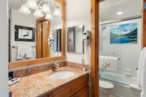 Un baño con lavamanos de encimera de granito, un espejo con una obra de arte de ciervo, un secador de pelo y una zona de ducha con mampara de vidrio.