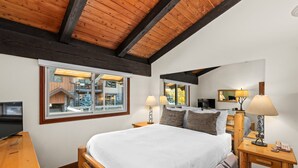 A bedroom with a wooden bed, two bedside tables with lamps, a TV, and a window with a view of a building.