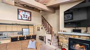 Amplia sala de estar con chimenea, cocina equipada con electrodomésticos modernos, una escalera y una camiseta deportiva grande enmarcada en la pared.