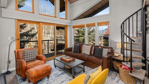 A spacious living room with a brown leather sofa, a yellow armchair, and a coffee table. There is a staircase with a black railing and a view of the outdoors through large windows.