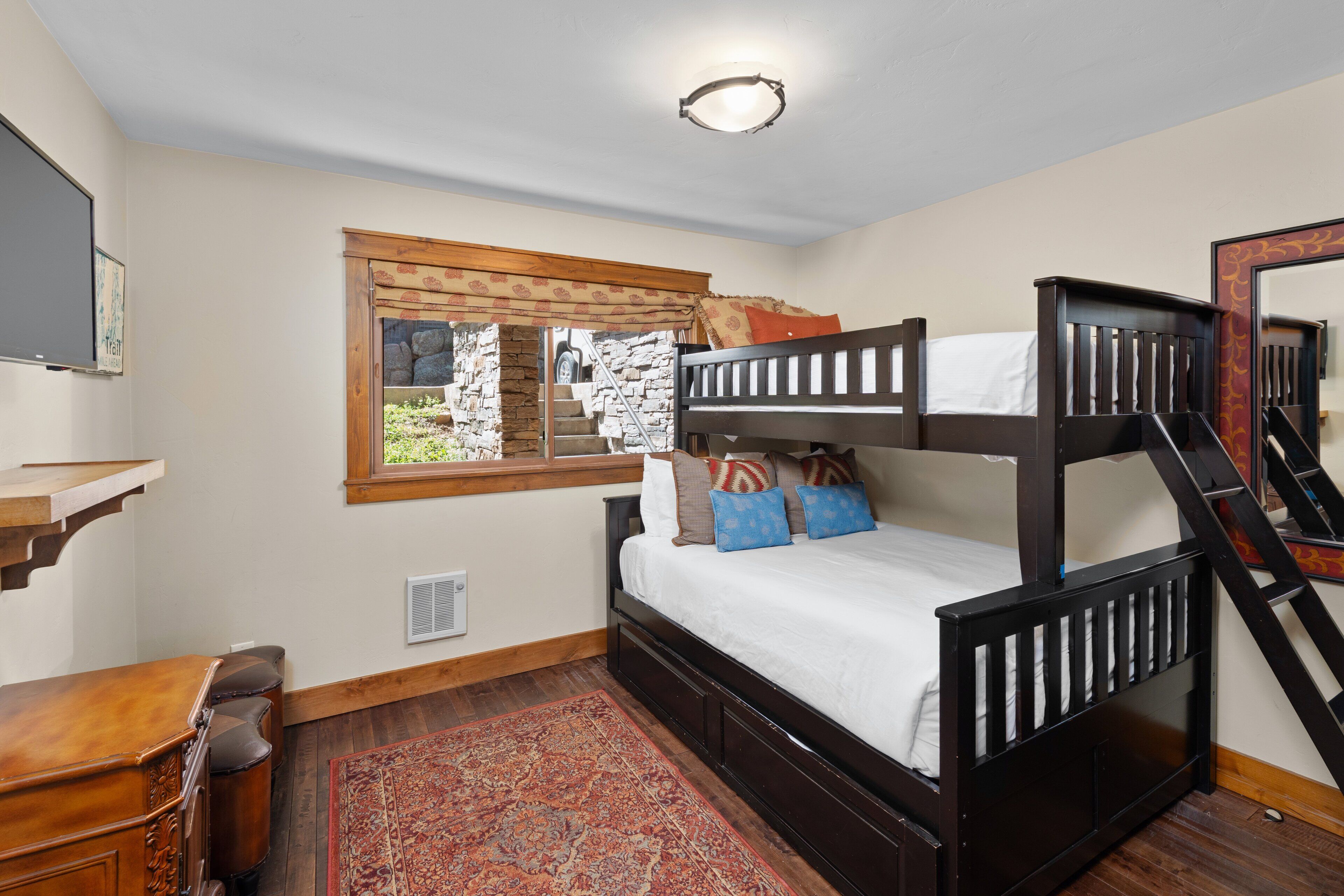A bunk bed with a wooden frame, a window with a view of a stone wall and greenery, a red patterned rug, and a wooden desk with a chair.