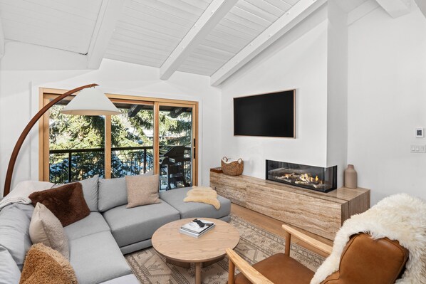 A modern living room with a grey sectional sofa, a round wooden coffee table, a TV mounted on the wall, and a fireplace. - Crestwood 1114 Premier 3 Bedroom plus Loft (Snowmass Village)