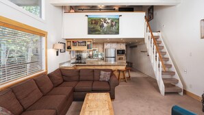 Un vaste espace de vie comprend un canapé d’angle marron, une table basse en bois et un fauteuil bleu. On aperçoit à l’arrière-plan une cuisine avec un réfrigérateur en acier inoxydable et un coin repas avec une table et des chaises en bois. Un escalier mène à un niveau supérieur où se trouve un grand tableau.