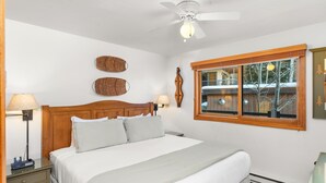 A bedroom with a large bed, wooden headboard, two wall-mounted lamps, a ceiling fan, and a window with a view of trees.