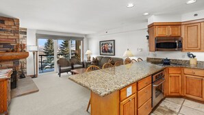 A spacious kitchen with granite countertops, wooden cabinets, and a large island. There is a dining area with wicker chairs and a view of trees through glass doors.