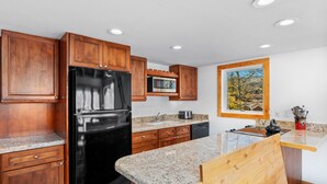 Une cuisine moderne avec des meubles en bois, des plans de travail en granit, un réfrigérateur noir, un four à micro-ondes et une fenêtre donnant sur les arbres.