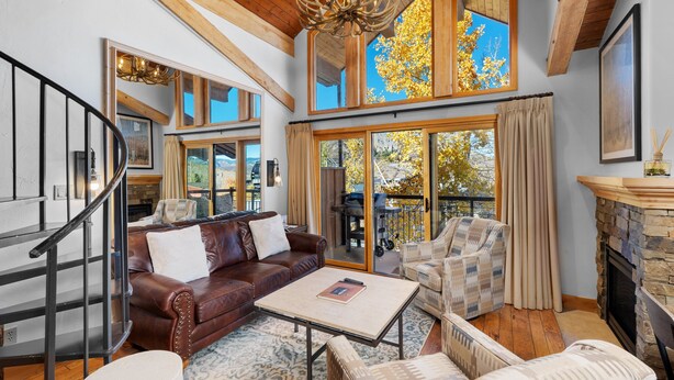 Ein geräumiges Wohnzimmer mit einer braunen Ledersofa, gemusterten Sesseln, einem Couchtisch und einem Kamin. Eine Treppe mit Metallgeländer ist links zu sehen. Große Fenster bieten Blick ins Freie.