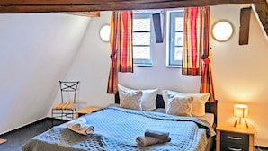 Ein gemütliches Schlafzimmer mit schrägem Dach, einem Bett mit blauer Decke, zwei Kissen, einem Sessel und einem Nachttisch mit Lampe.