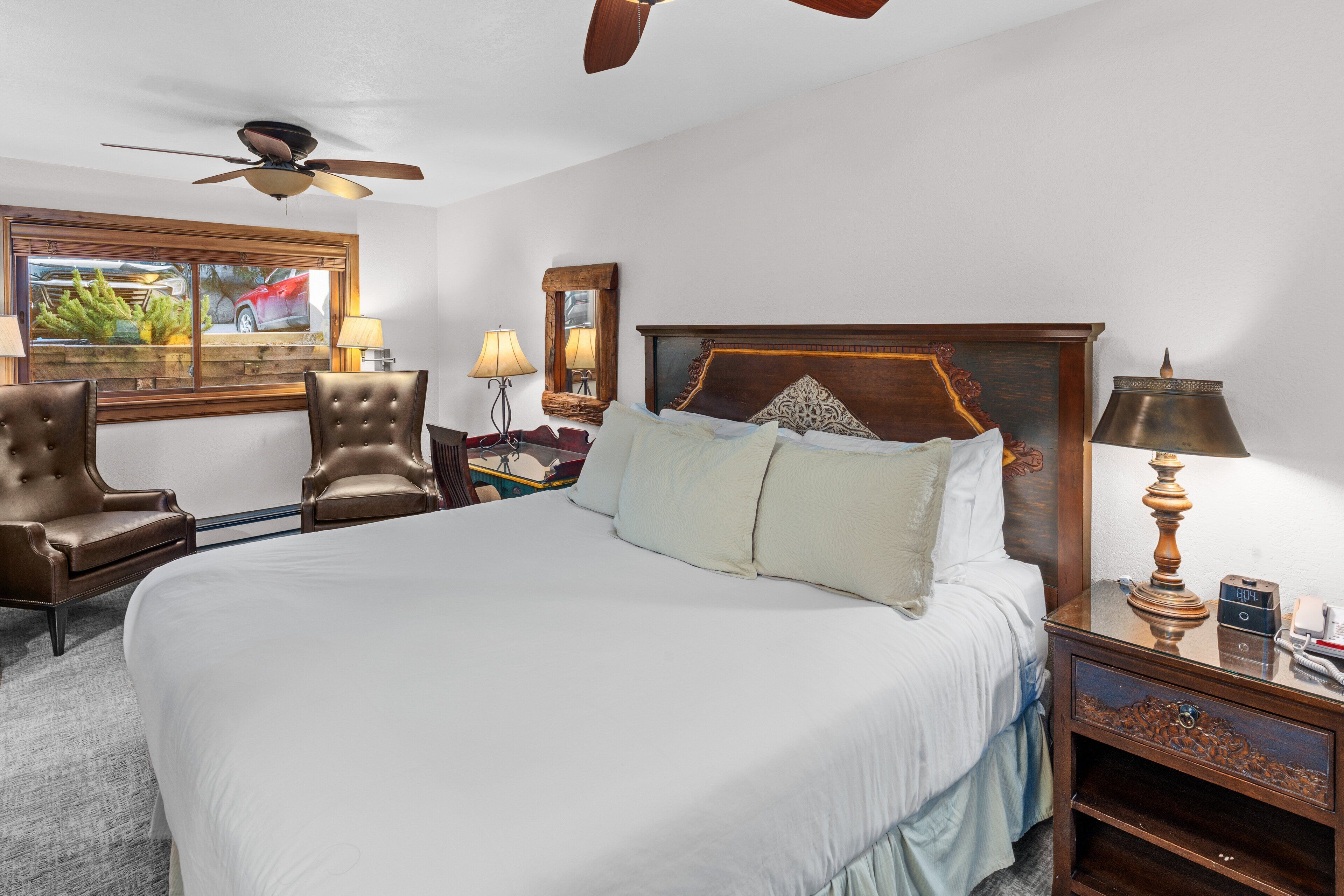 A hotel room with a large bed, two chairs, a nightstand with a lamp, a ceiling fan, and a window with a view of a red car.