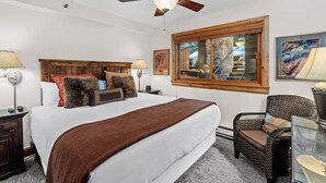 A bedroom with a large bed, a wooden headboard, a ceiling fan, a window with a view, and a wicker chair. - Crestwood 2109 Premier 2 Bedroom (Snowmass Village)