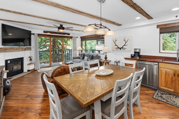 A spacious living area with a wooden dining table, white chairs, a fireplace, and a large TV.