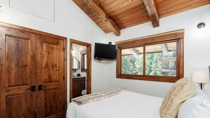 Un dormitorio con techo de madera, una cama con ropa de cama blanca, un televisor montado en la pared y una ventana con vistas a árboles.