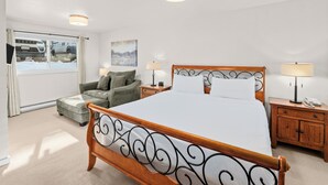 A bedroom with a large bed, a wooden nightstand, a chair, a lamp, and a window with a view of a parked car.