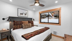 A bedroom with a bed, a nightstand, a ceiling fan, a window with a view of a vehicle, and a framed picture on the wall.