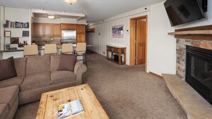 A living room with a sectional sofa, a coffee table, a fireplace, and a kitchen area with wooden cabinets and a flat-screen TV. - Crestwood 2117 Standard 2 Bedroom (Snowmass Village)