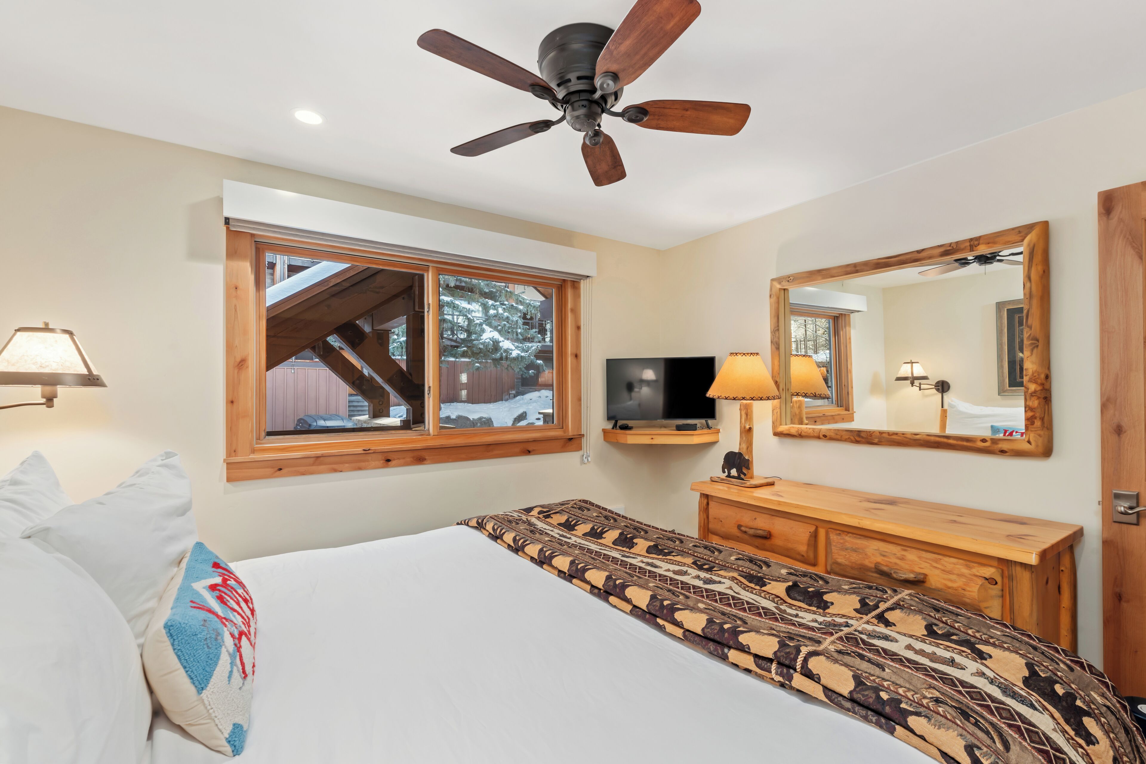 Un dormitorio con una cama, ventilador de techo, televisor, cómoda de madera, espejo y ventana con vistas a un paisaje nevado.