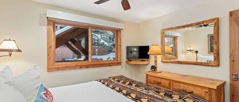 Un dormitorio con una cama, ventilador de techo, televisor, cómoda de madera, espejo y ventana con vistas a un paisaje nevado.