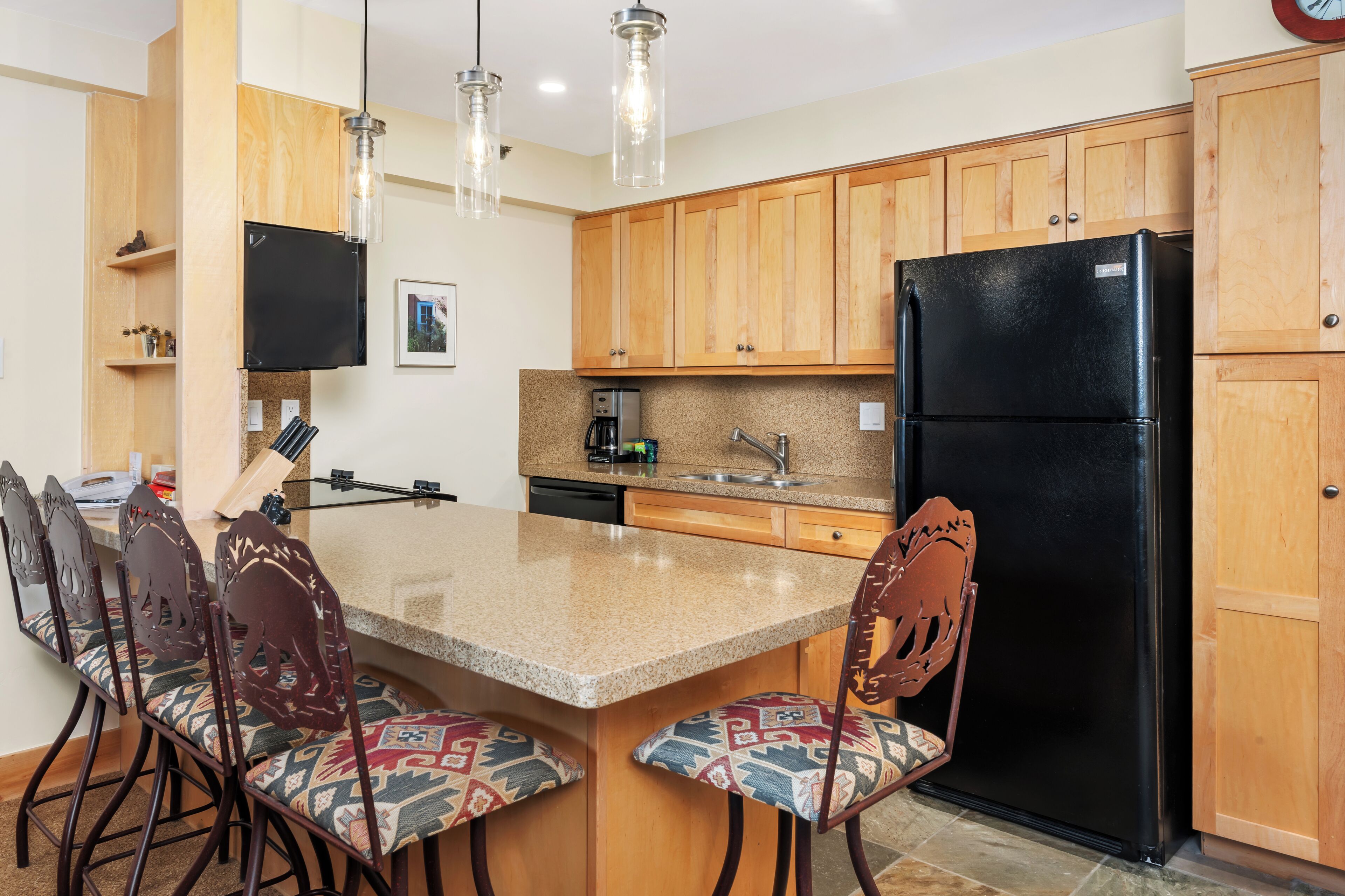 Una cocina con armarios de madera, un refrigerador negro, encimera de granito y un comedor con sillas estampadas.