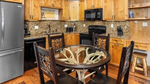 A kitchen with wooden cabinets, a stainless steel refrigerator, a granite countertop, a glass-top dining table, and a black stove.