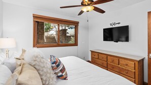 A bedroom with a bed, a wooden dresser, a television on the wall, and a window with a view of the outside.