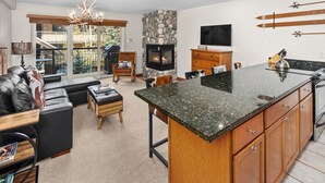 A modern kitchen with a stone fireplace, a dining area, and a living room with a sofa and a chandelier.