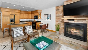 A modern kitchen with wooden cabinets, a dining area with chairs, a flat-screen TV mounted above a fireplace, and a green tray table with magazines.